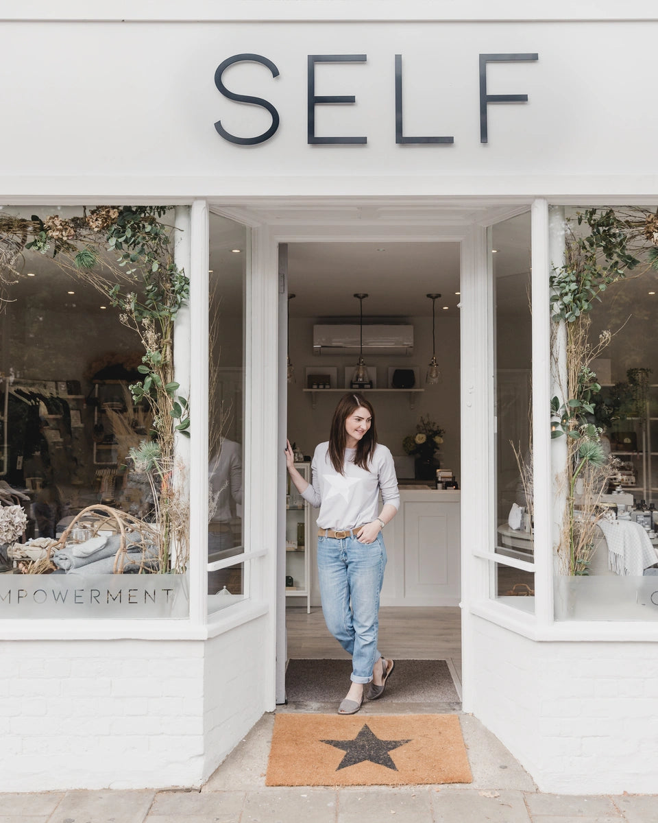Suzie, owner of SELF, posed in the doorway of SELF in Woodhall Spa, looking down and off to the right with one hand in her pocket and the SELF sign above her head