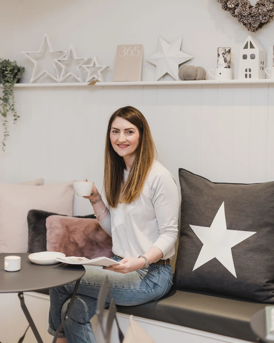 Suzie, owner of SELF, sat on bench seating with a cup of coffee in hand smiling towards the camera, holding a lifestyle magazine