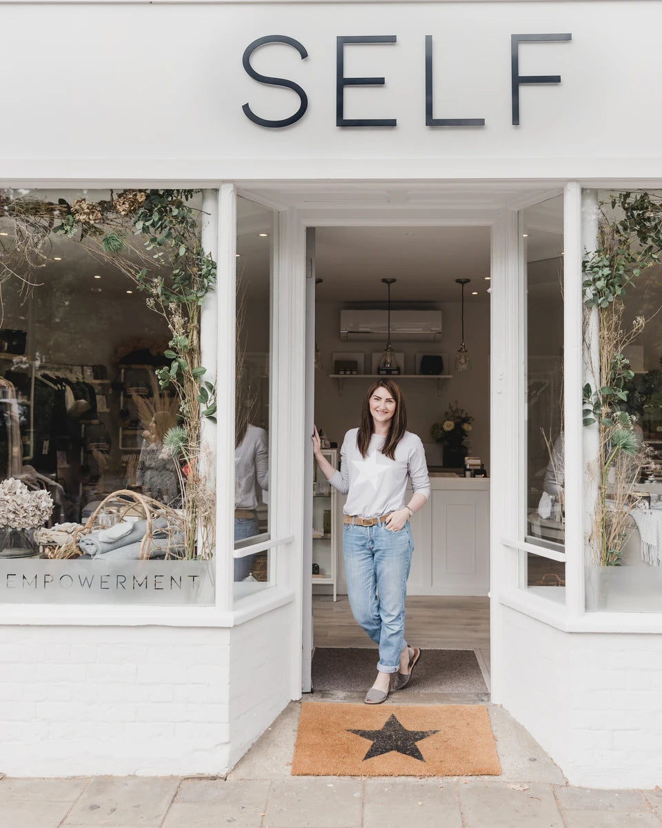 Suzie, owner of SELF, sat on bench seating with a cup of coffee in hand smiling towards the camera, holding a lifestyle magazine