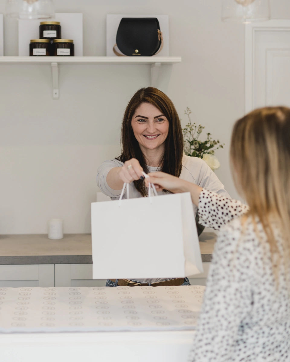 Our owner, Suzie, stood smiling behind the grey counter in one of our SELF boutiques. A customer is in the foreground with a white gift bag being handed to them by Suzie