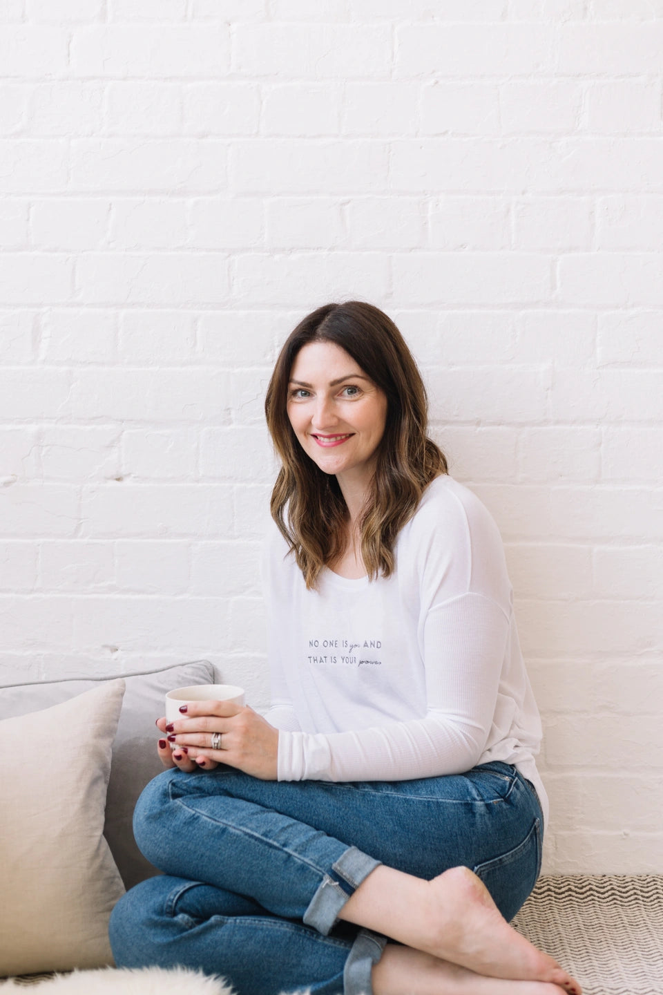 Suzie, owner of SELF, sat on the floor with her legs folded to the side holding a warm mug of tea and smiling at the camera