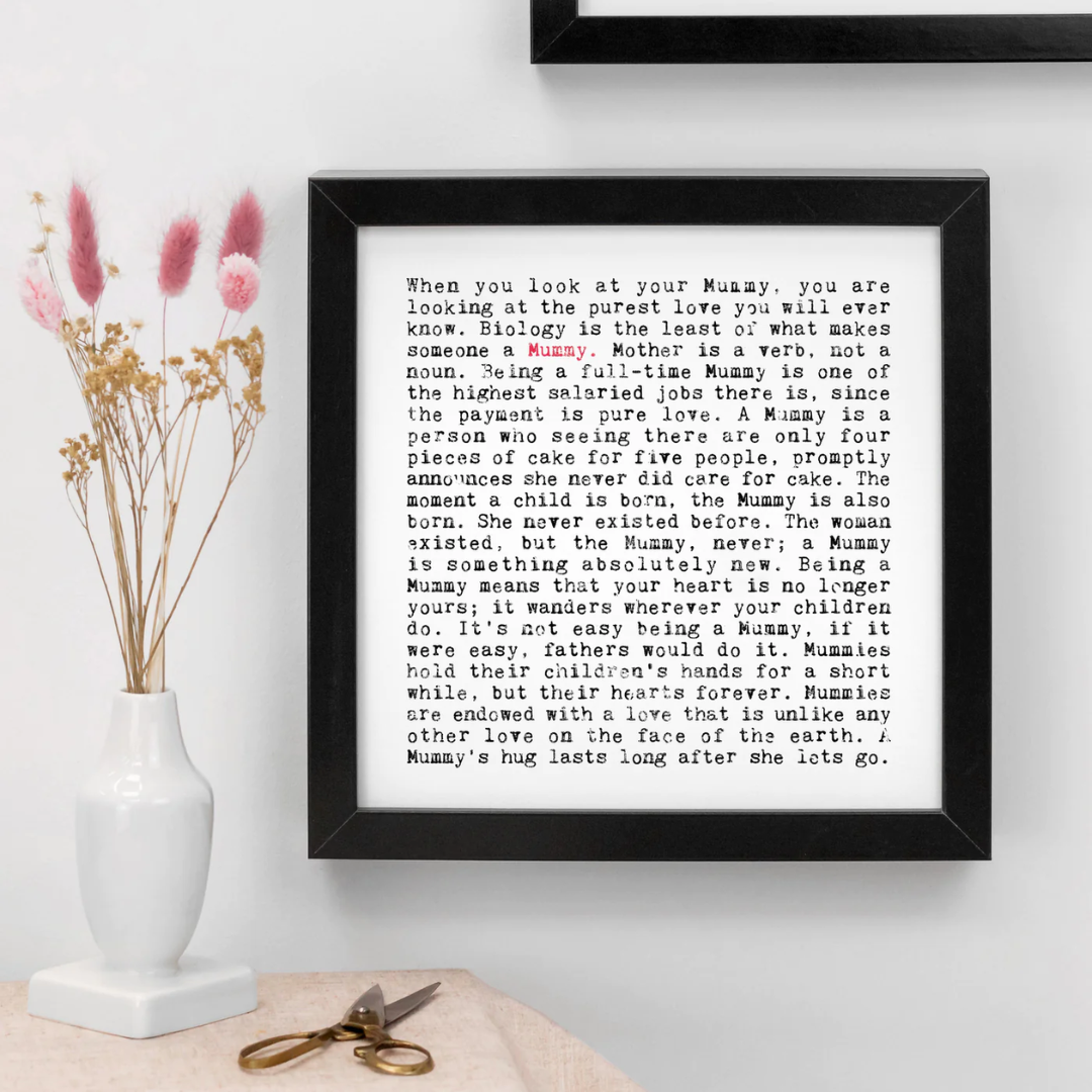 A typography 'mum' inspired print hung on a white wall with a black frame. A small white vase is next to the print with dried pink flowers inside.